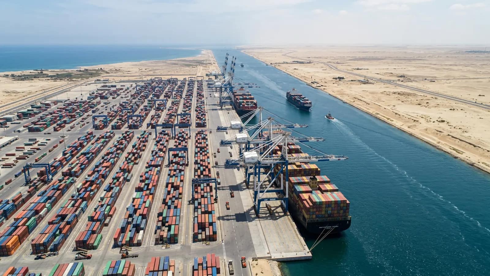 Container terminal on a canal bank, cranes working a container ship, with flat desert-coloured coast in the distance